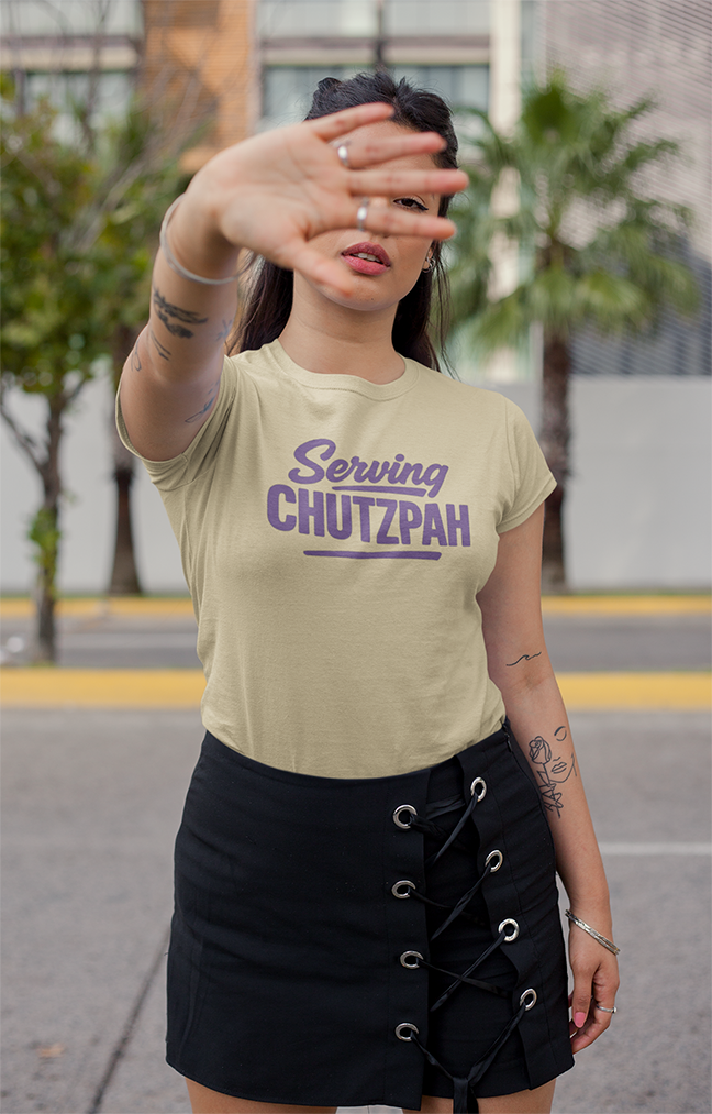 Woman wearing a beige t-shirt with 'Serving Chutzpah' text, standing outdoors.