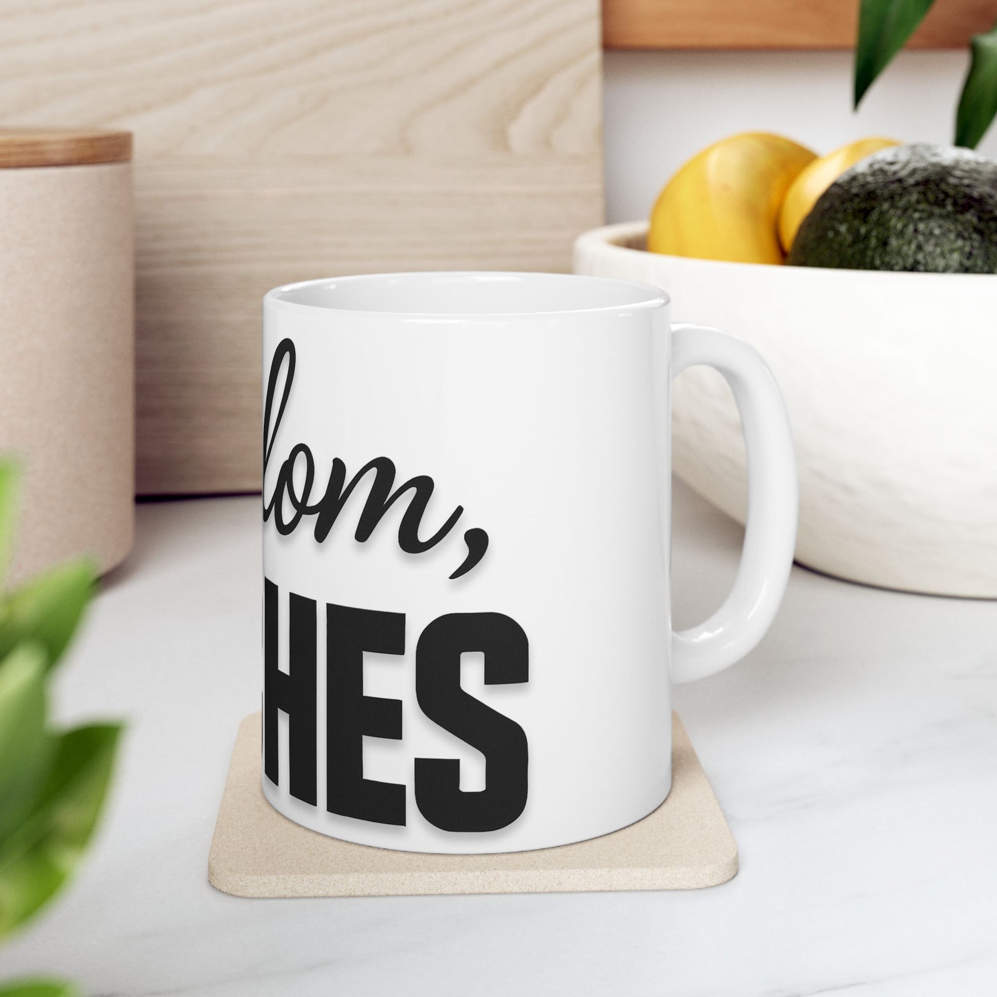 White mug with black text 'shalom, bitches' on a coaster, placed on a surface with a plant and container in the background.