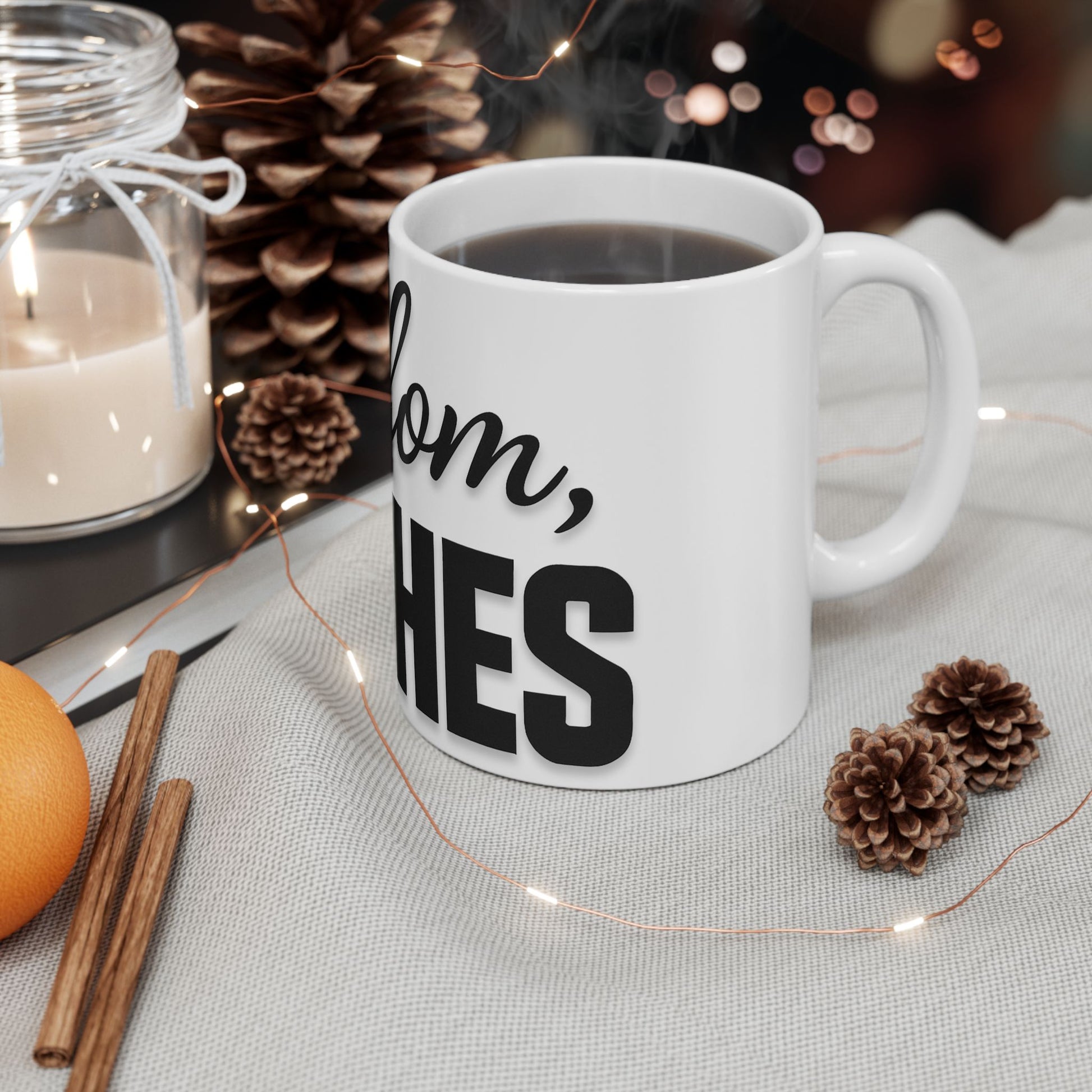 White mug with text 'shalom, bitches', candle, cinnamon sticks, and pine cones on a table.