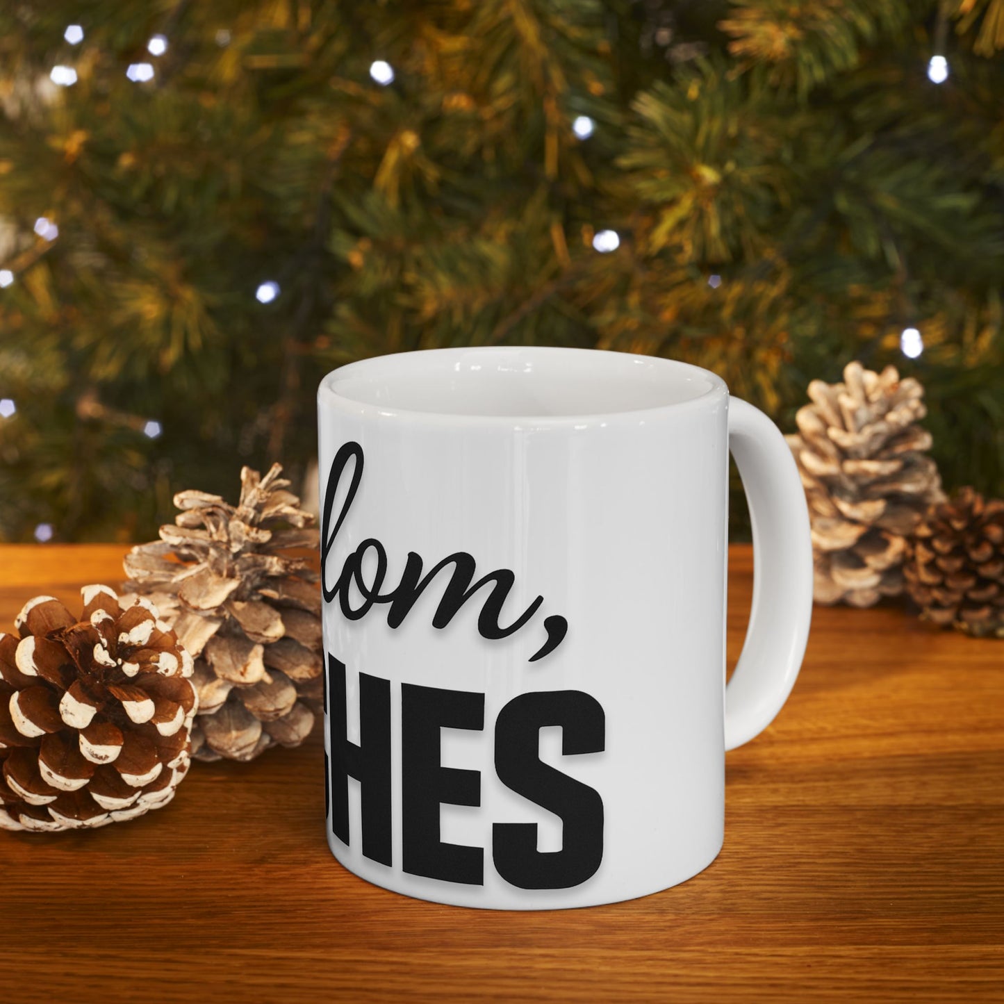 White mug with text 'shalom, bitches' in front of a Christmas tree and pine cones.