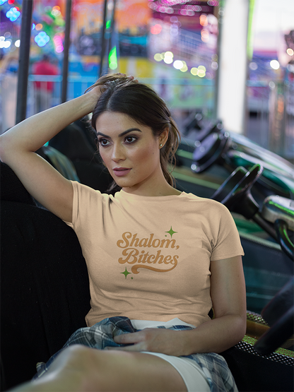 Woman wearing a beige 'shalom bitches' t-shirt with text in an amusement park setting