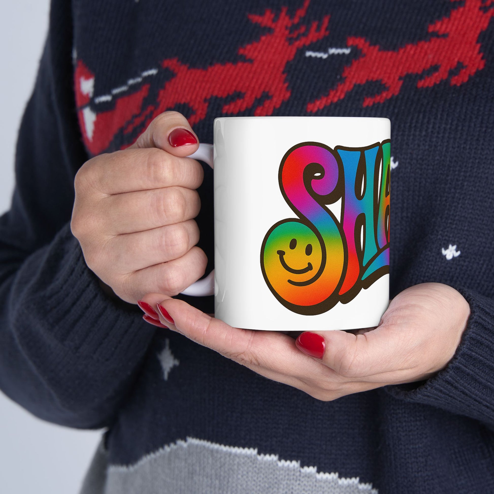 Person holding a mug with a colorful 'Shalom' design, wearing a dark sweater with red deer patterns.