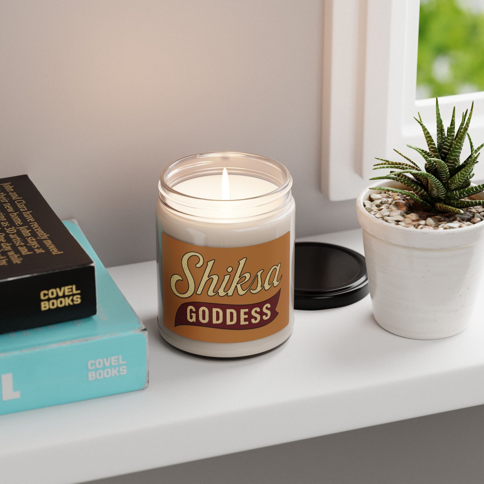 Candle labeled 'Shiksa Goddess' on a shelf with books and a plant
