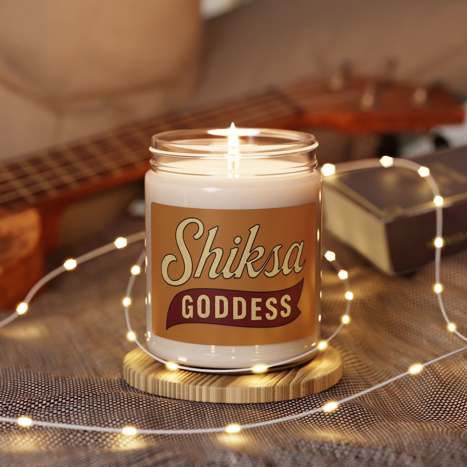 Candle in a jar labeled 'Shiksa Goddess' with a guitar and string lights in the background