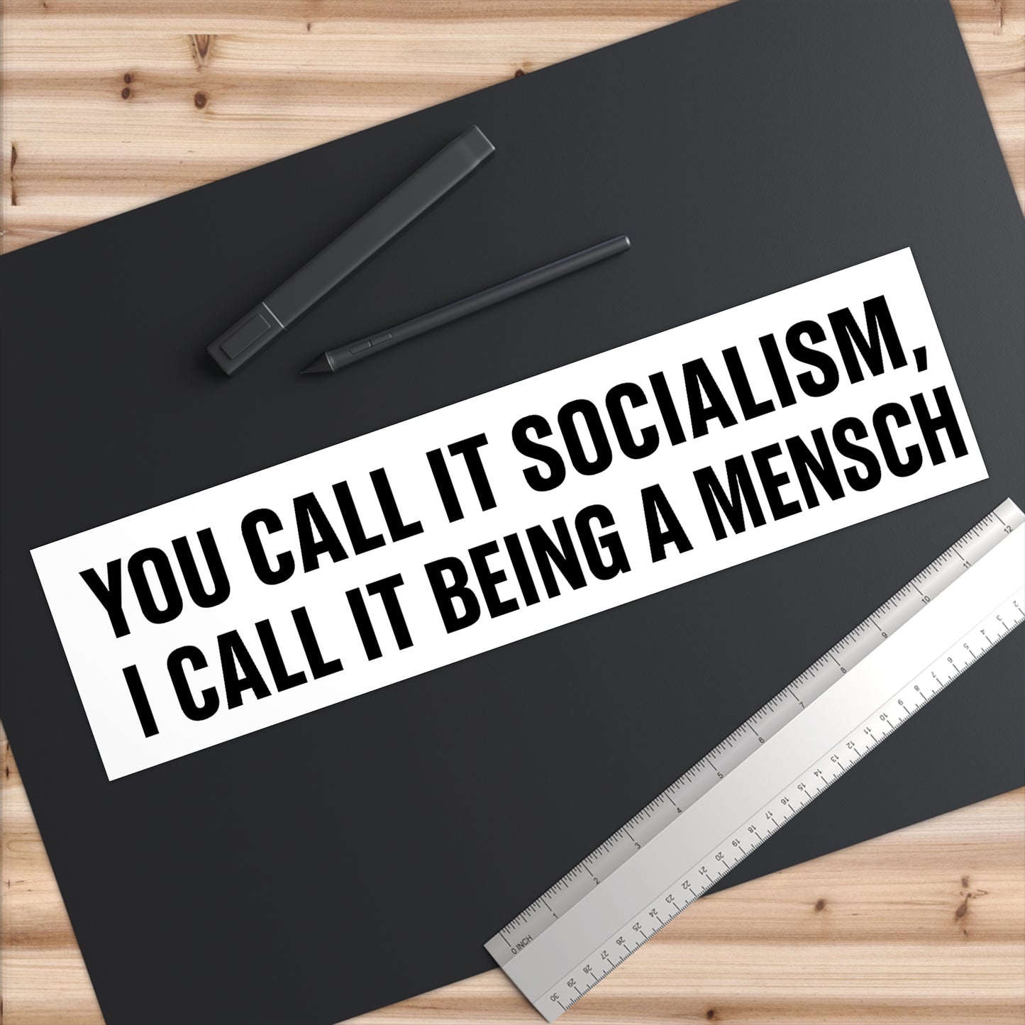 Black mat with text 'You call it socialism, I call it being a mensch' on a wooden surface with pens and a ruler.