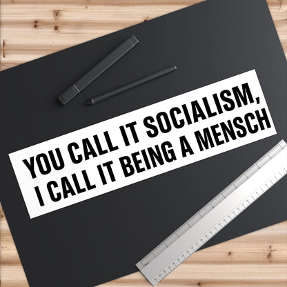 Black mat with text 'You call it socialism, I call it being a mensch' on a wooden surface with pens and a ruler.