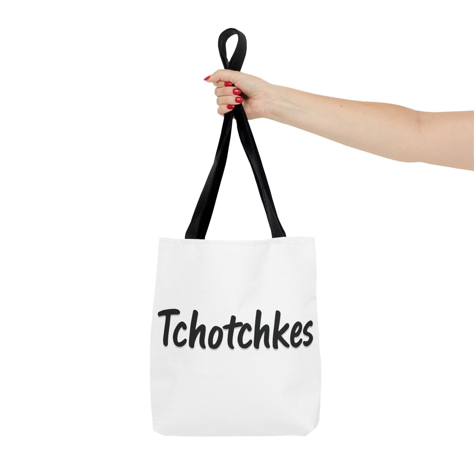 White tote bag with 'Tchotchkes' text held by a hand with red nail polish on a white background