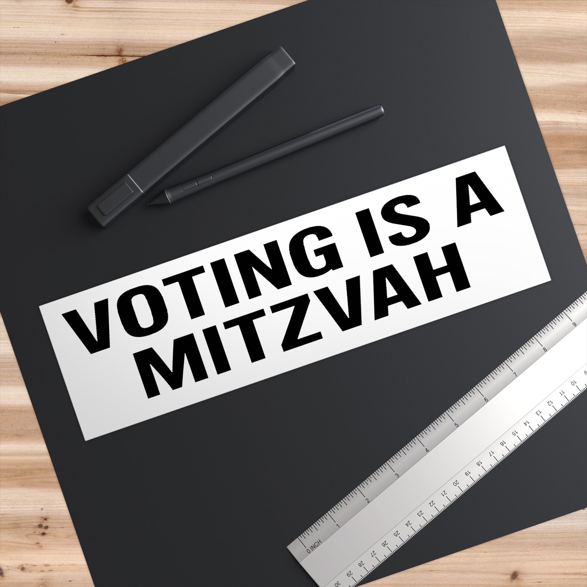Black paper with 'Voting is a Mitzvah' text on a wooden surface with pens and a ruler.