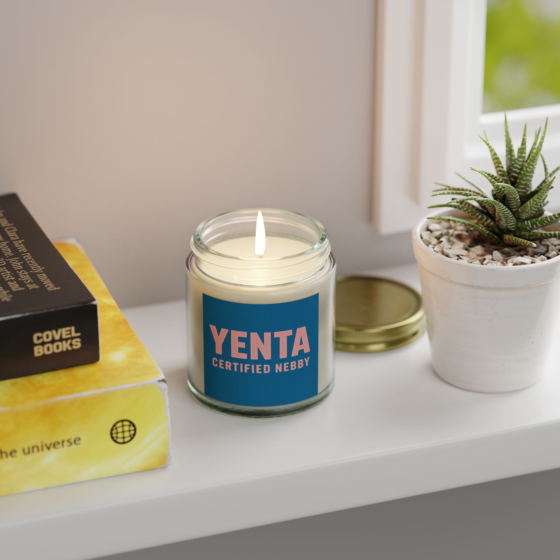 Candle with a 'Yenta Certified Nebby' label on a shelf with books and a plant.
