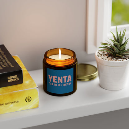 Candle with 'Yenta Certified Nebby' label on a shelf with books and a plant